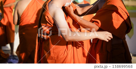 A senior monk adjusts a novice robe as hands tighten the woven belt around orange cloth in sunlight with focus on texture folds and precise fitting. 138699568