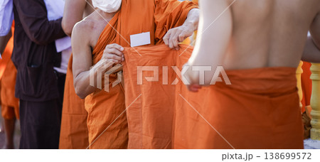 A senior monk adjusts a novice robe as hands tighten the woven belt around orange cloth in sunlight with focus on texture folds and precise fitting. A senior monk adjusts a novice robe as hands tighten the woven belt around orange cloth in sunlight with focus on texture folds and precise fitting. 138699572