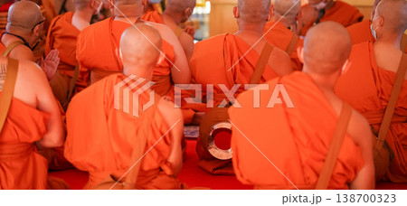New monks sit in a large group inside the hall during ordination. Orange robes and warm temple light fill the frame. New monks sit in a large group inside the hall during ordination. Orange robes and warm temple light fill the frame. 138700323