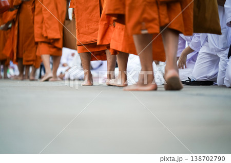Barefoot monks in orange robes walk past kneeling people in white. The low angle highlights feet, fabric and the ritual path. 138702790