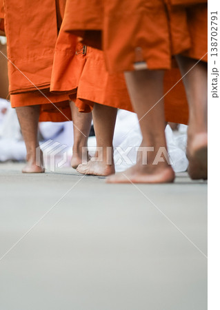 Barefoot monks in orange robes walk past kneeling people in white. The low angle highlights feet, fabric and the ritual path. 138702791