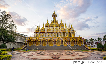 An ornate Thai temple stands centered beneath a cloudy evening sky. Gold details and wide steps define the grand facade. 138702792