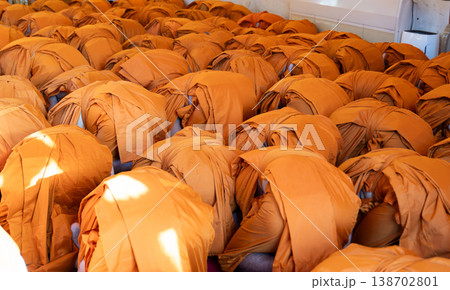 Monks bow forward together in a packed indoor ritual. Orange robes cover the scene in ordered rows. 138702801