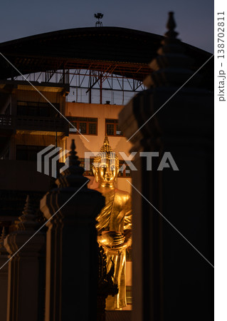 A golden standing Buddha glows between dark temple pillars at dusk. Warm light shapes the statue against the shadowed background. 138702811