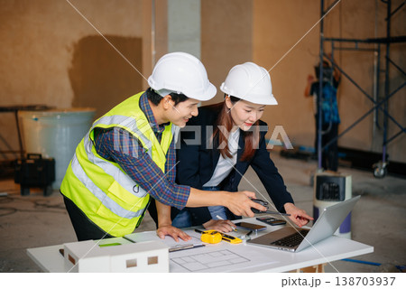 Asian architects at construction site using laptop and blueprints for project planning. Wearing hard hats and vests, they discuss design 138703937