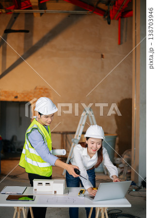 Asian engineers at building site reviewing architecture model. Wearing hard hats and safety vests, they discuss layout, planning, teamwork 138703946
