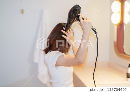 Young woman drying long hair with a hair dryer while looking at the mirror after a shower in the bathroom at home or hotel room during vacation. Hairstyles and daily morning routine Young woman drying long hair with a hair dryer while looking at the mirror after a shower in the bathroom at home or hotel room during vacation. Hairstyles and daily morning routine 138704495