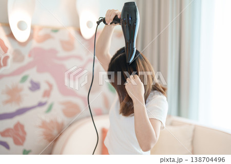Young woman drying long hair with a hair dryer after a shower in home or hotel room during vacation. Hairstyles and daily morning routine Young woman drying long hair with a hair dryer after a shower in home or hotel room during vacation. Hairstyles and daily morning routine 138704496