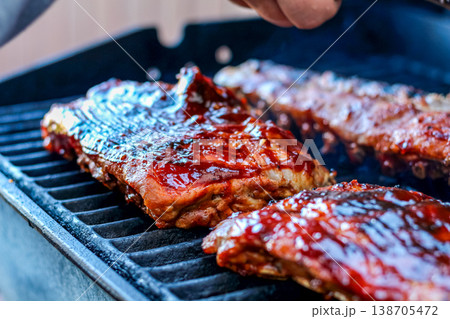 Grilled pork ribs coated with barbecue sauce on a barbecue grill, with smoke rising and a hand applying sauce in the background Grilled pork ribs coated with barbecue sauce on a barbecue grill, with smoke rising and a hand applying sauce in the background 138705472