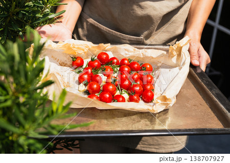 Hands holding a baking tray with fresh cherry tomatoes rosemary and garlic on parchment paper. 138707927