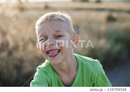 Happy boy with blond hair and a green T-shirt, smiling broadly and sticking his tongue out. Happy childhood. Summer holidays. 138709506