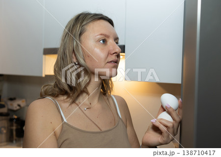 Woman holding two fresh eggs in modern kitchen near refrigerator. Morning breakfast preparation and healthy eating routine concept for daily nutrition and family meals. Woman holding two fresh eggs in modern kitchen near refrigerator. Morning breakfast preparation and healthy eating routine concept for daily nutrition and family meals. 138710417