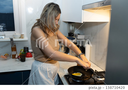 Woman frying egg on stove in modern kitchen. Morning breakfast preparation and daily routine. Fresh home cooking concept for healthy start to day. 138710420