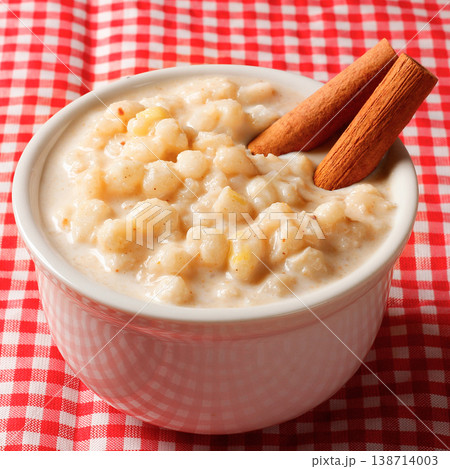 Canjica or mungunza creme, a traditional Brazilian dessert made with white corn, served in a white bowl with cinnamon sticks on a red checkered tablecloth 138714003