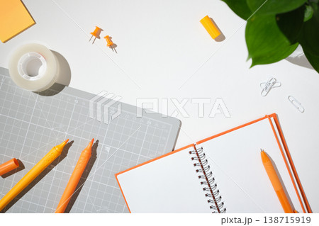 Orange stationery and crafting tools on a white background. Open blank notebook, markers, cutting mat, tape, and push pins with copy space. 138715919