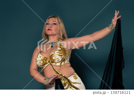 Studio portrait of confident middle aged blonde woman belly dancing. Female dancer wears an elaborate white and golden costume and holds black veil, posing gracefully against teal background Studio portrait of confident middle aged blonde woman belly dancing. Female dancer wears an elaborate white and golden costume and holds black veil, posing gracefully against teal background 138716121