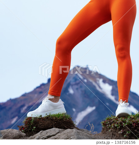 Low angle view of a fit woman's legs in bright orange leggings and white sneakers. She stands on a rocky summit with a snowy mountain in the background. Active lifestyle and travel concept Low angle view of a fit woman's legs in bright orange leggings and white sneakers. She stands on a rocky summit with a snowy mountain in the background. Active lifestyle and travel concept 138716156