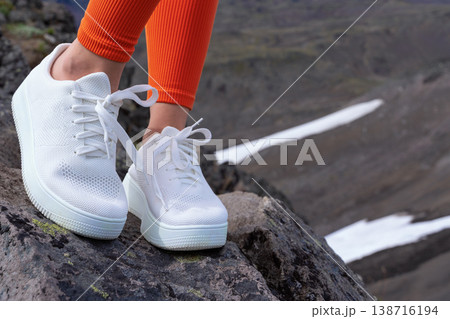 Closeup of a woman in white sneakers and bright orange leggings on a rocky mountain. The modern footwear contrasts with the rugged natural landscape. Active lifestyle, travel, and adventure concept Closeup of a woman in white sneakers and bright orange leggings on a rocky mountain. The modern footwear contrasts with the rugged natural landscape. Active lifestyle, travel, and adventure concept 138716194