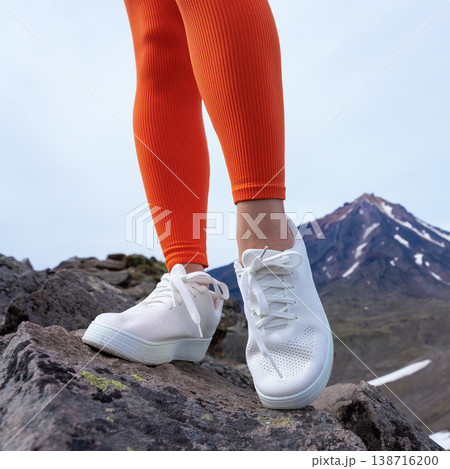 Low angle close up of a woman's feet in white sneakers and vibrant orange leggings. She is standing on dark volcanic rocks with a snow-capped mountain in the background. Active lifestyle concept Low angle close up of a woman's feet in white sneakers and vibrant orange leggings. She is standing on dark volcanic rocks with a snow-capped mountain in the background. Active lifestyle concept 138716200