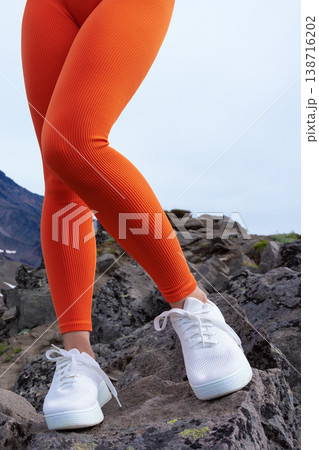 Low-angle view of a woman's legs in vibrant orange ribbed leggings white sneakers. Athlete standing on rugged rocks, perfect for active lifestyle, fitness, outdoor fashion concepts. Bright colors pop 138716202