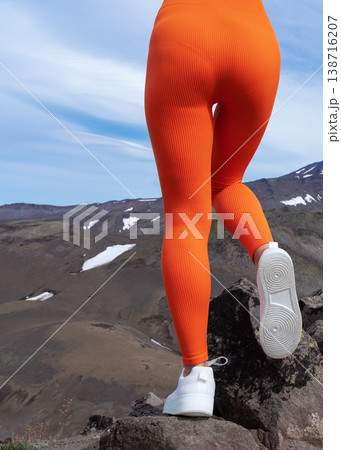 Low angle view of a fit woman's legs in vibrant orange ribbed leggings and white sneakers. Woman stands on a rocky summit, exploring a rugged mountain valley. Active lifestyle and travel concept 138716207