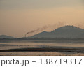 Serene landscape at dawn with factory chimney emitting smoke against backdrop of hills and calm body of water 138719317