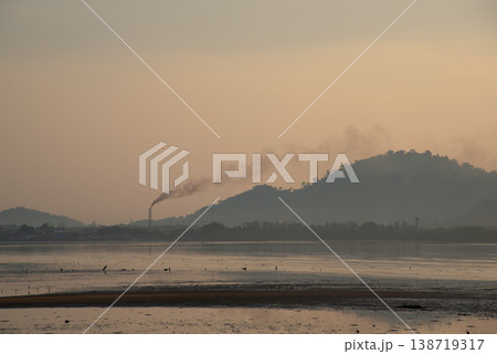 Serene landscape at dawn with factory chimney emitting smoke against backdrop of hills and calm body of water Serene landscape at dawn with factory chimney emitting smoke against backdrop of hills and calm body of water 138719317