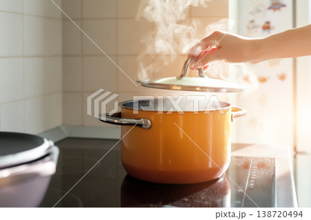 Female hand open lid of enamel steel cooking pan on electric hob with boiling water or soup and scenic vapor steam backlit by warm sunlight at kitchen. Kitchenware utensil and tool at home background 138720494