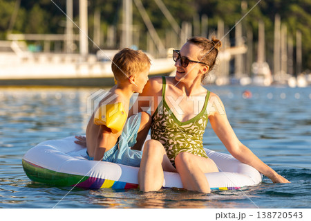 Mother and young boy sit inflatable raft calm sea water marina warm sunset light. Summer vacation moment shows family bonding, happiness, parenting care, childhood joy, and relaxed travel lifestyle Mother and young boy sit inflatable raft calm sea water marina warm sunset light. Summer vacation moment shows family bonding, happiness, parenting care, childhood joy, and relaxed travel lifestyle 138720543