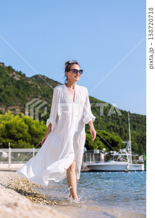 Portrait smiling woman wear white dress walk barefoot pebble sand beach sea bright summer day sunshine Stylish female person enjoy vacation relaxation lifestyle natural beauty freedom coastal scenery 138720718