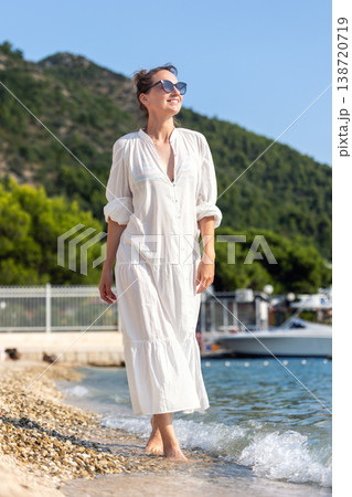 Portrait smiling woman wear white dress walk barefoot pebble sand beach sea bright summer day sunshine Stylish female person enjoy vacation relaxation lifestyle natural beauty freedom coastal scenery 138720719