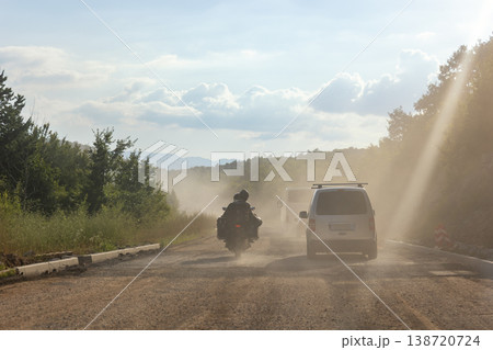 Dusty road construction traffic motorcycle rider van driving unpaved highway rural landscape sunlight rays.. Transport scene heavy traffic dirt surface haze infrastructure repair rural landscape 138720724