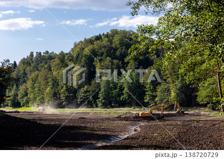 Excavators deepen clean drained Trakoscan reservoir basin by forested hills clear blue sky. Earthmoving machines reshape muddy bottom channels during landscape restoration and water management works Excavators deepen clean drained Trakoscan reservoir basin by forested hills clear blue sky. Earthmoving machines reshape muddy bottom channels during landscape restoration and water management works 138720729