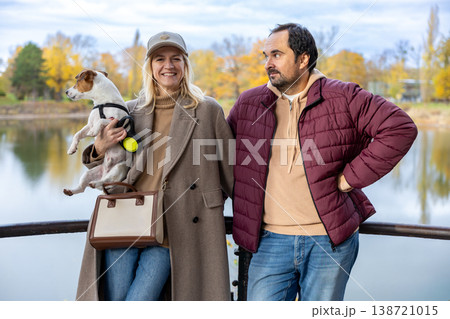 Happy Adult Couple With Small Dog Standing Near Autumn Lake Outdoor Portrait 138721015