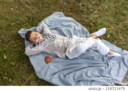 Young boy relaxing on a soft blanket in the park with a fresh red apple beside him. Young boy relaxing on a soft blanket in the park with a fresh red apple beside him. 138721479