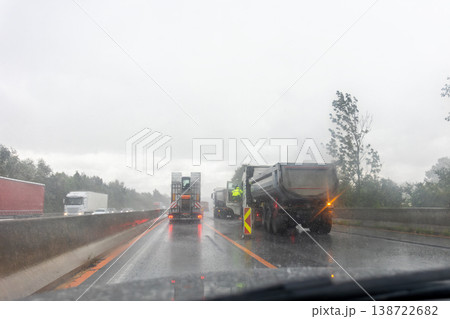 Driver POV rain highway roadworks trucks workers wear safety gear traffic poor visibility wet asphalt conditions. Vehicles move slowly construction site zone warning signs barriers reflective lights Driver POV rain highway roadworks trucks workers wear safety gear traffic poor visibility wet asphalt conditions. Vehicles move slowly construction site zone warning signs barriers reflective lights 138722682