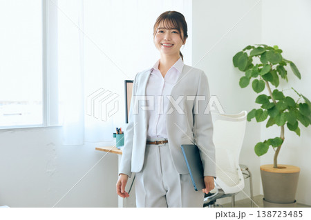 オフィスでファイルを持ち笑顔で立つライトグレーのスーツ姿の若い日本人女性ビジネスパーソン オフィスでファイルを持ち笑顔で立つライトグレーのスーツ姿の若い日本人女性ビジネスパーソン 138723485