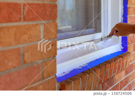 Person applies white paint to window frame while preparing area with blue tape 138724500