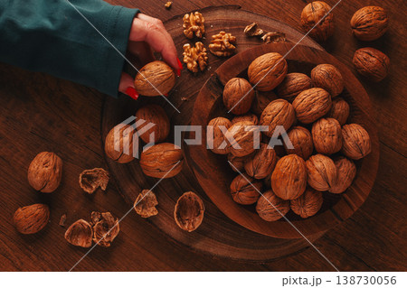 a woman's hand taking a walnut from a wooden bowl filled with walnuts. There are shells and walnuts scattered around. 138730056