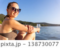 Smiling woman in sunglasses relaxes near calm water holding glass during warm sunset. Natural beauty, summer vacation mood, peaceful landscape freedom leisure serene travel atmosphere by quiet shore 138730077