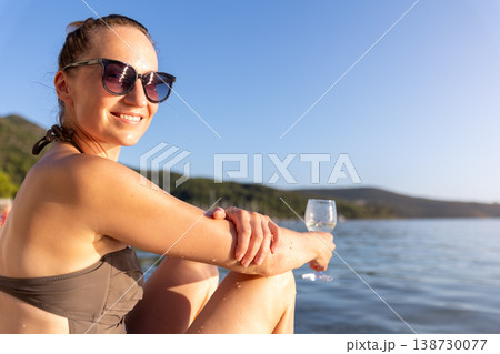 Smiling woman in sunglasses relaxes near calm water holding glass during warm sunset. Natural beauty, summer vacation mood, peaceful landscape freedom leisure serene travel atmosphere by quiet shore Smiling woman in sunglasses relaxes near calm water holding glass during warm sunset. Natural beauty, summer vacation mood, peaceful landscape freedom leisure serene travel atmosphere by quiet shore 138730077