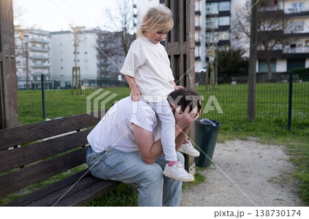 Funny 4 year old girl climbing on her tired father trying to rest on a bench Funny 4 year old girl climbing on her tired father trying to rest on a bench 138730174