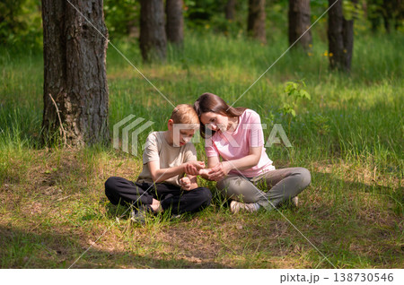 mother and son are sitting side by side in a forest clearing and looking at something in their palms mother and son are sitting side by side in a forest clearing and looking at something in their palms 138730546