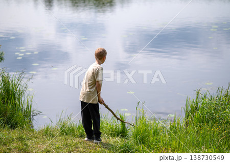 Boy standing on lakeside with a long stick, playing near the water in nature on a summer day Boy standing on lakeside with a long stick, playing near the water in nature on a summer day 138730549