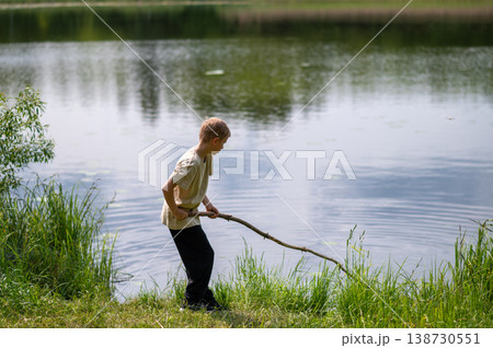 Boy standing on lakeside with a long stick, playing near the water in nature on a summer day Boy standing on lakeside with a long stick, playing near the water in nature on a summer day 138730551