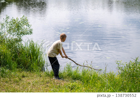 Boy standing on lakeside with a long stick, playing near the water in nature on a summer day Boy standing on lakeside with a long stick, playing near the water in nature on a summer day 138730552