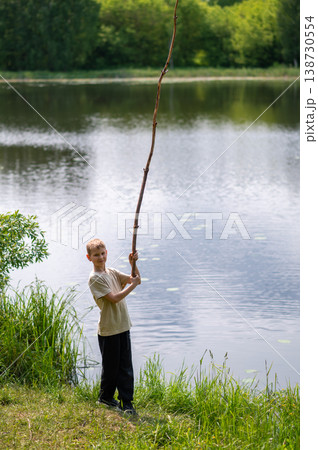 Boy standing on lakeside with a long stick, playing near the water in nature on a summer day Boy standing on lakeside with a long stick, playing near the water in nature on a summer day 138730554