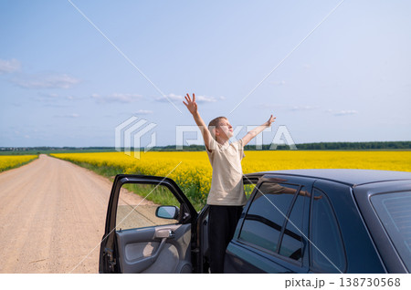 Happy boy standing with raised arms on country road in rapeseed field near car on sunny summer day, joyful outdoor adventure Happy boy standing with raised arms on country road in rapeseed field near car on sunny summer day, joyful outdoor adventure 138730568