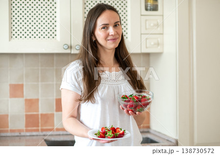 Smiling woman holding bowl and plate of fresh strawberries in kitchen, promoting healthy lifestyle 138730572
