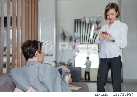 Businesswomen, one standing and one sitting, discussing notes together in stylish modern workspace. Discussion, notes, businesswomen, teamwork, office. Businesswomen, one standing and one sitting, discussing notes together in stylish modern workspace. Discussion, notes, businesswomen, teamwork, office. 138730984